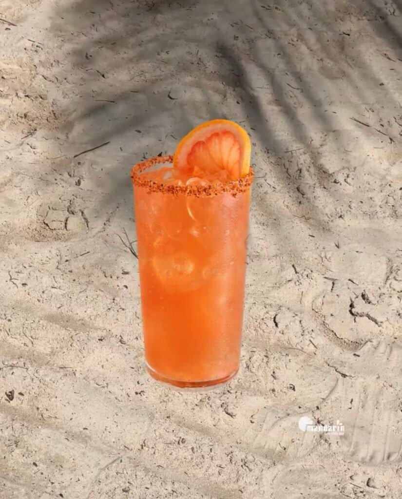 MANDARIN SEASIDE BAR Cocktail τοποθετημένο στην άμμο μπροστά από τη θάλασσα, με καλοκαιρινή ατμόσφαιρα.